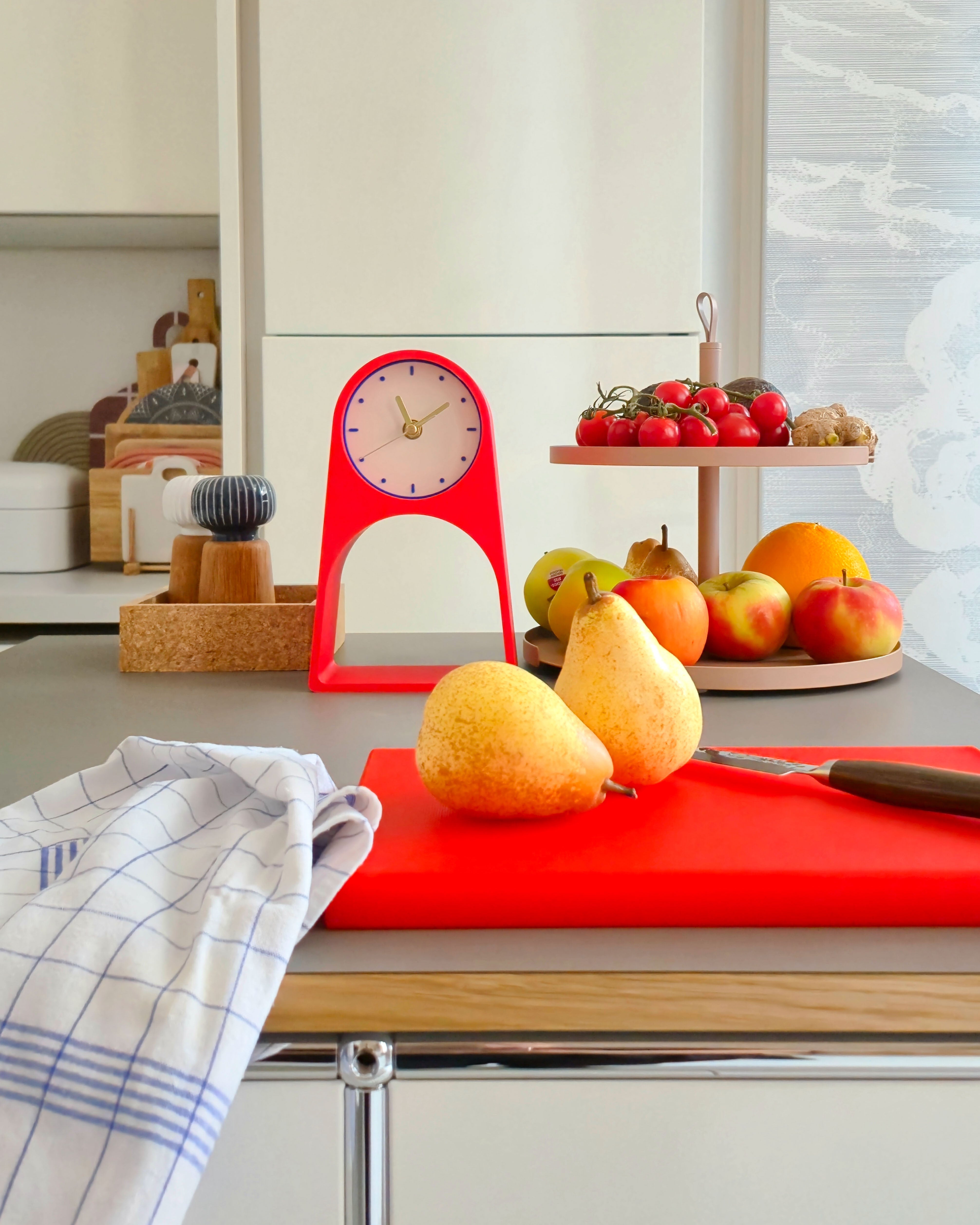 Red Table Clock LEIF standing on a kitchen cupboard with fruits around it.